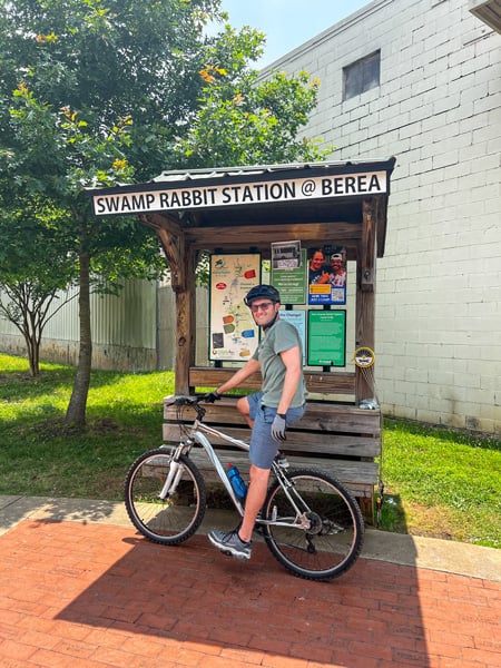 Tom on bike in front of sign Swamp Rabbit Station At Berea information board