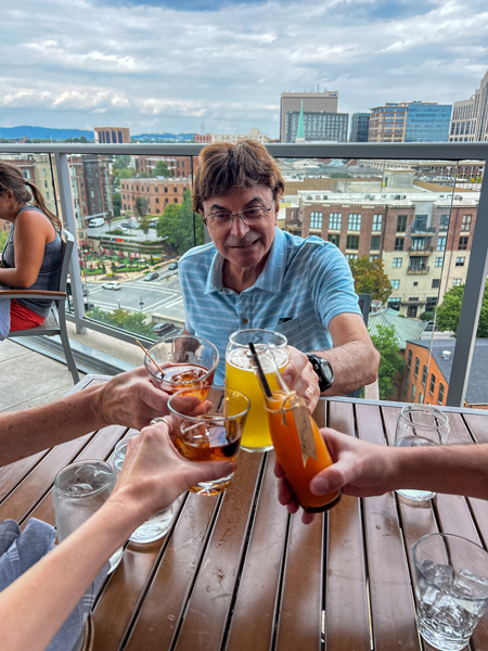 Christine's family doing a cheers with their drinks at Up on the Roof in Greenville, SC; pictured with their hands is her dad, a white brunette male with view of Downtown Greenville from above behind him