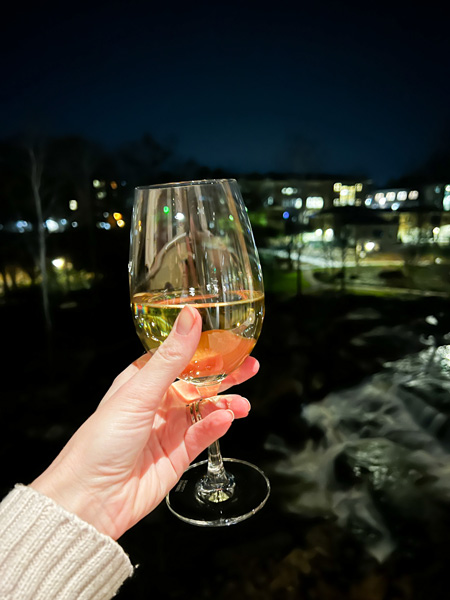 Christine's white hand in a beige sweater holding up a white wine glass over Falls Park on the Reedy at the Grand Bohemian Lodge Greenville from their Lobby Bar 